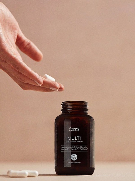 A clean, high-end studio shot of a Daily Nutrient Support supplement bottle paired with a hand placing a capsule, using soft neutral tones to convey purity and quality.